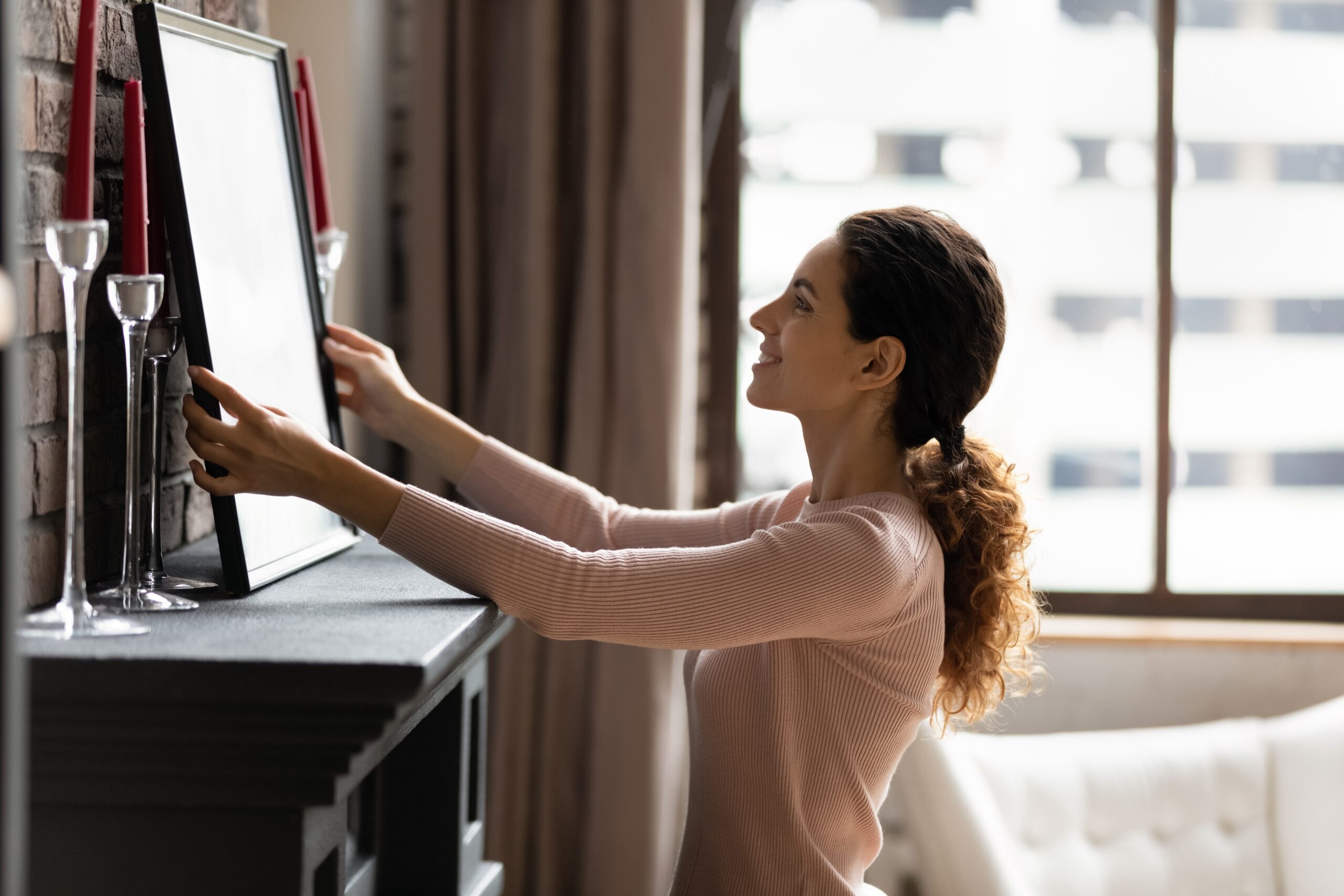 A smiling woman with her hair pulled back in a curly ponytail places a framed picture on a dark mantelpiece. She is wearing a light pink long-sleeved shirt. Two tall, clear glass candle holders with long red candles flank the picture. A large window with a view of city buildings is in the background.